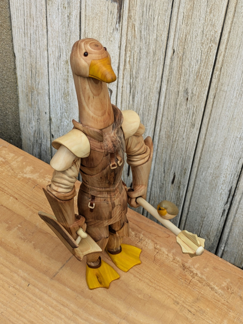 Woodcarved humanoid goose wearing plate armour. She's holding a shield and a flanged mace. On the handle of the mace, a red robin landed. The eyes of the goose are made of glass. The feet and the beak are painted in burnt yellow, as the breast of the red robin. The upper part of the robin is painted in brown/grey  and its eyes and beak in black.