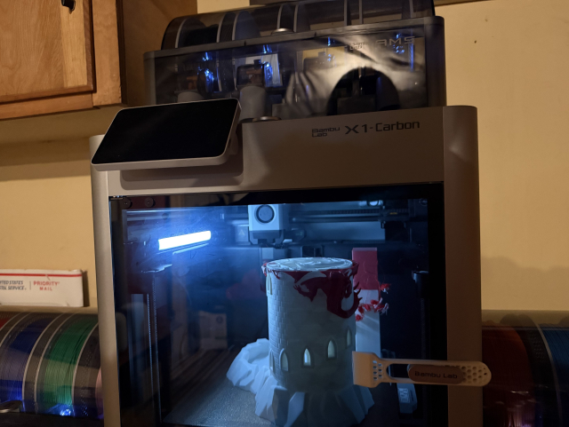 A Bambu Lab X1-Carbon is actively printing a cylindrical structure with small arched windows, resembling a tower or castle turret. The top of the object has some red filament details. The printer has a digital display screen on the top left corner, and there is a tool labeled "Bambu Lab" attached to the front of the printer. In the background, there are spools of filament in various colors, including green and red, visible on a shelf. The setting appears to be a workshop or a room with wooden cabinets.