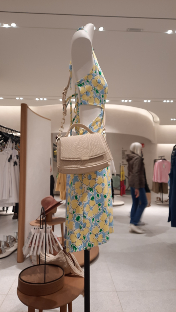 A mannequin with a yellow-and-blue summer dress, halter-necked and with cut-outs at the waist. A small handbag hanging off one shoulder is about twice the width of the whole supposed 'woman's body'.  Now follows the fuller context and scene description, the same as in the other photo in this toot, so you can skip if already heard: A mannequin in the women's clothing section of a department store. Beyond, clothes on racks and a few distant customers. The mannequin purports to be a woman's body. It has no head, of course. The 'body' is barely centimetres wide, a stick figure, a tall pencil of a 'woman'. The white mannequin has no shape or curves. It is we-are-worried-about-your-health skinny, but supposed to be inspirational and aspirational. They are designing women's mainstream clothing to fit unrealistically skinny bodies like this.