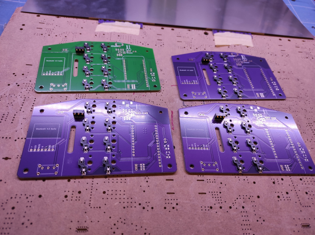Vier Platinen mit Bauteilen für die Verbindung mit Tasten und Akku. Eine Platine ist grün, drei sind violett. Den Untergrund bildet eine Holzplatte mit vielen Bohrlöchern. Im Hintergrund liegt eine Metallplatte auf einer Schneidmatte.