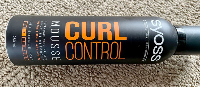 A bottle of curl control mousse by Syoss lying on a textured surface.