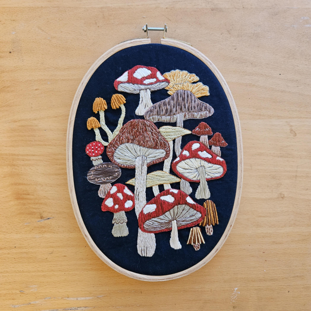 An oval embroidery hoop containing a mushroom themed design, depicting a diverse array of fungi on a navy blue background. 