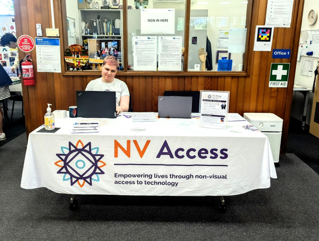 Sascha sitting at a table with NV Access tablecloth and fliers, with laptop ready to demonstrate NVDA