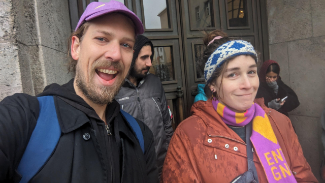 two people smiling in front of a concrete government building with several people threatened with eviction behind them. everybody looks cold and wet