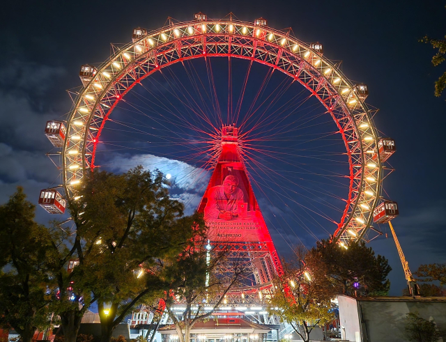 A photo of the Viennese Giant Ferris Wheel at night, its lights are on and the full Moon is illuminating the clouds in the background.