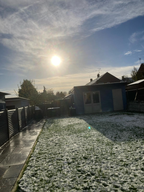 A snow-dusted garden on a sunny day with a clear blue sky. Sun glare is visible. Snow covers grass and parts of a paved path. A blue shed with sunlit windows stands in the background, flanked by a wooden fence. Bare trees and rooftops are also visible in the distance.