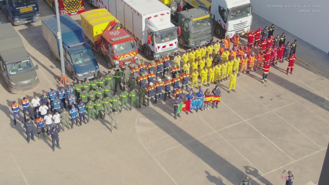 A large assembly of rescue teams and emergency responders lined up for a group photo. The teams are wearing uniforms of various colors, representing different organizations and nations. Flags of Spain, Portugal, and the European Union are displayed prominently. Emergency vehicles, including trucks and ambulances, are parked in the background