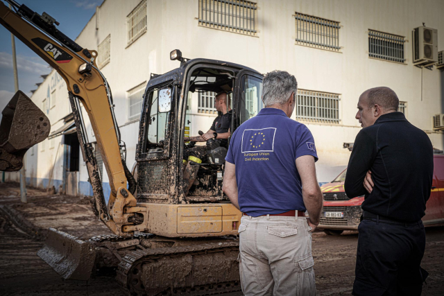 A civil protection worker in a blue European Union Civil Protection shirt observes a construction vehicle clearing debris. A firefighter operates the muddy excavator, and another person in black attire stands next to the observer, all engaged in post-disaster recovery.