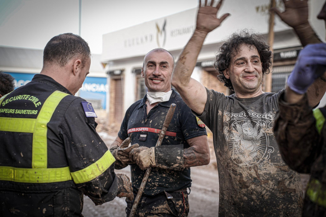 A group of rescuers and volunteers covered in mud during a recovery operation. One man is smiling, holding a shovel, while another has his hands raised in celebration. The uniforms indicate civil security personnel, with visible badges and reflective elements.