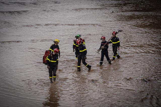 A group of rescue workers wearing helmets and reflective uniforms walking through muddy floodwaters. Some carry tools such as shovels, while others carry backpacks or rescue equipment. The water appears deep and turbulent, indicating recent flooding.