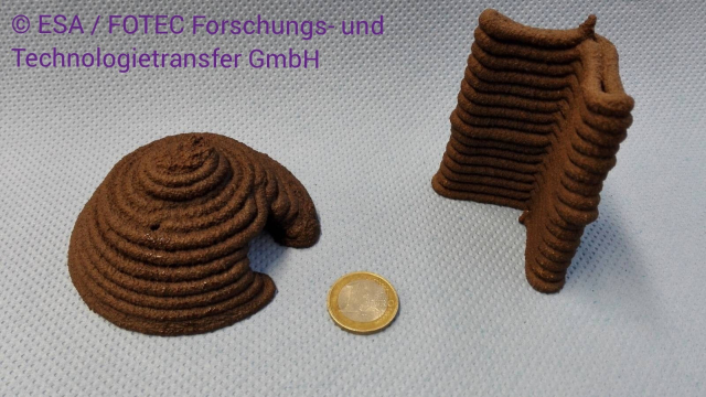 Auf dem Foto sind zwei braune 3D-gedruckte Objekte, links eine runde Hütte und rechts ein Eck das ein Wandteil sein könnte. In der Mitte liegt eine 1€-Münze um einen Größenvergleich zu haben. Links oben stehen die Credits: © ESA / FOTEC Forschungs- und Technologietransfer GmbH