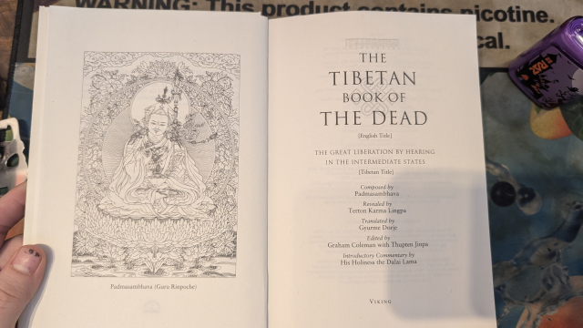 This image shows an open book with the title The Tibetan Book of the Dead. The left page features an intricate illustration of Padmasambhava (Guru Rinpoche), a significant figure in Tibetan Buddhism, seated in a meditative posture amidst floral motifs. The right page lists the title in English and its alternate titles, along with information about its composition, translation, and editorial contributions. It states the text was composed by Padmasambhava, revealed by Terton Karma Lingpa, translated by Gyurme Dorje, and edited by Graham Coleman with Thupten Jinpa, with an introductory commentary by His Holiness the Dalai Lama. The book focuses on teachings about the intermediate states (bardo) and liberation.