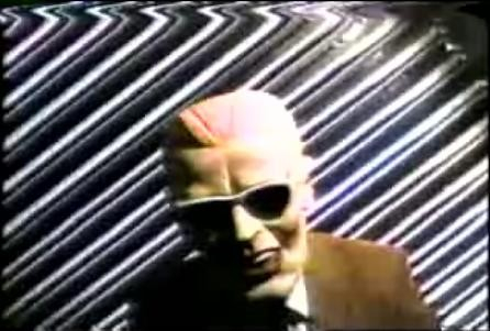 Low quality video screenshot of a person in a rubber mask and sunglasses in front of a diagonal pattern. 