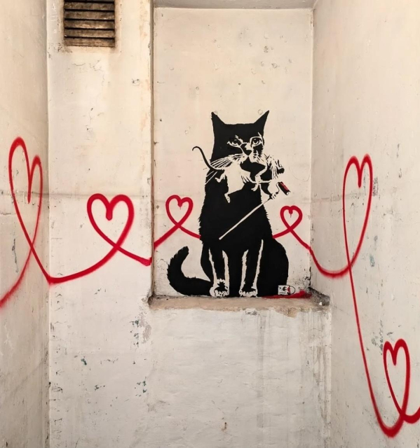 Streetartwall.  A stencil mural with a cat, a rat and red hearts was sprayed into a white wall niche. The black cat by artist "Tabby" sits in the niche and has a rat in the style of "Banksy" in its mouth. The Banksy rat is holding a stick with a paint roller. From the cat, curved and interconnected hearts extend beyond the two walls into another room of an exhibition.
Info: The TABBY Vienna exhibition at Hotel Kunst BTQ will take place from November 29 to December 1. 2024.
Tabby has freely designed the walls of the exhibition rooms. Admission is free but only open via guest list in the Website.