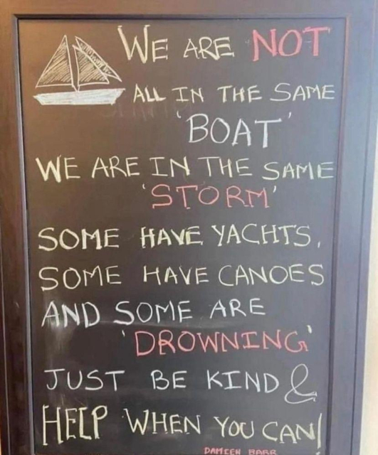 A chalkboard sign with a hand-drawn sailboat at the top displays the following message in colorful chalk: "We are NOT all in the same 'BOAT'. We are in the same 'STORM'. Some have yachts, some have canoes, and some are 'DROWNING'. Just be kind & HELP when you can!" The words "NOT," "STORM," and "DROWNING" are highlighted in red, while the rest of the text alternates between white and yellow. The sign is framed in wood, conveying a meaningful reminder about empathy and kindness.