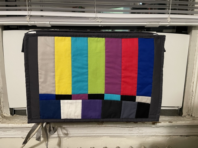 A fabric quilt covers a window-installed air conditioner. The quilt is made up of the characteristic coloured rectangles of a TV test pattern.