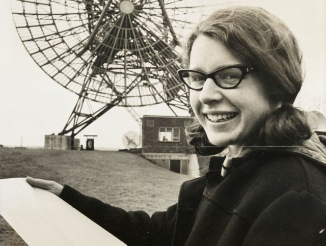 Jocelyn Bell Burnell in the 1960s, in front of a radio telescope. she is a white woman with dark hair, wearing a sensible duffle coat given how cold it must have been out there.

