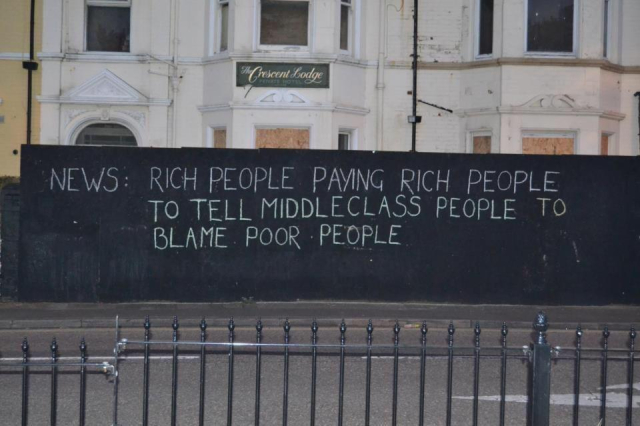 Street art on a black wall outside a boarded-up building with the text: 'NEWS: RICH PEOPLE PAYING RICH PEOPLE TO TELL MIDDLECLASS PEOPLE TO BLAME POOR PEOPLE.' The graffiti, written in simple white chalk-style lettering, critiques societal and economic dynamics. The location appears to be in an urban area, with a fence in the foreground and a sign reading 'The Crescent Lodge' visible on the building behind the wall.