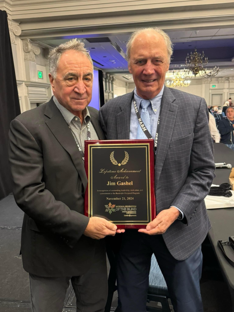 This image shows two senior gentlemen at what appears to be an awards ceremony or formal event. They are holding a Lifetime Achievement Award plaque presented to Jim Gashel on November 21, 2024. The plaque has a black face with gold lettering and appears to be mounted on a burgundy-colored wood base.  The setting is indoors in what looks like a formal event space or banquet hall, with decorative white molding visible in the background and what appears to be a chandelier. There's also a hint of blue lighting in the background.  Both men are dressed formally - one in a dark suit with a checked shirt, and the other in a gray plaid sport coat with a light blue dress shirt and tie.  - Made with PiccyBot