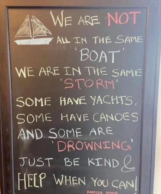 A chalkboard sign with a hand-drawn sailboat at the top displays the following message in colorful chalk: "We are NOT all in the same 'BOAT'. We are in the same 'STORM'. Some have yachts, some have canoes, and some are 'DROWNING'. Just be kind & HELP when you can!" The words "NOT," "STORM," and "DROWNING" are highlighted in red, while the rest of the text alternates between white and yellow. The sign is framed in wood, conveying a meaningful reminder about empathy and kindness.