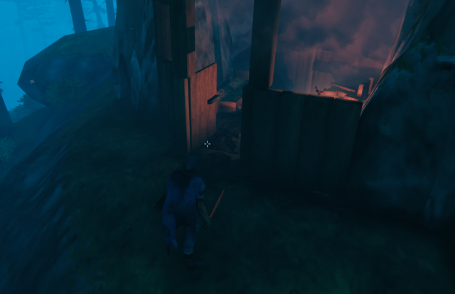 A hooded man stands before a cave mouth that has had wooden walls, furniture, and a campfire nstalled.