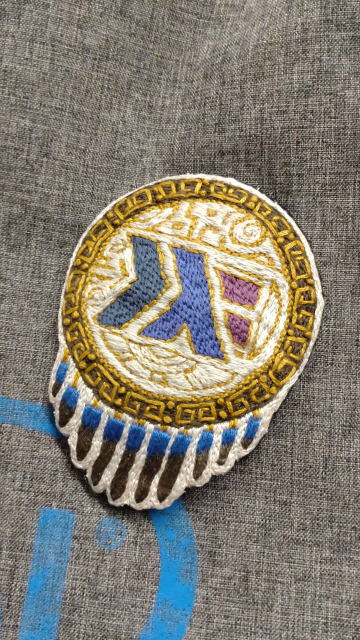 Una insignia bordada con un logotipo estilizado de Haskell con elementos azules, morados y dorados, sobre un fondo gris.