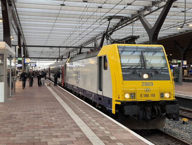 A silver locomotive with a yellow front and number 2869 and also E 186 119 in front of a rake of silver carriages waiting at a platform of Rotterdam Centraal.