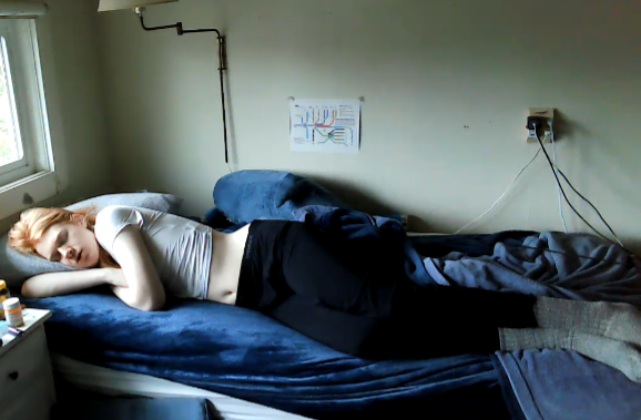 Peaceful sleeping red head facing the camera after sunrise showing a cute belly button and pinchable sides. Clothed in a white crop top and black shorts. Blahaj in the background.