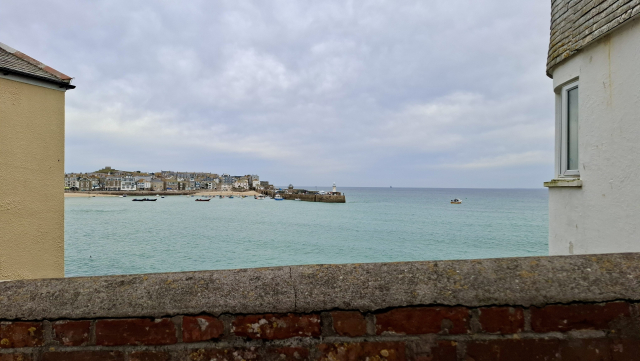 Peering over a low sloping old stone pier wall that borders the bottom of the scene. We can see the town of St. Ives in Cornwall across a bay of almost tropically light green sea below a light blue sky. A few boats are anchored in front of the town, which curves down to the water and a picturesque small white beacon on a jutting pier. Our view is framed on the left and right by the edges of two old stone houses, one yellow, one white with a corner window.  The feeling of the scene, to me: nothing dramatic, just simple and quiet, a glimpse of a pretty town as we near it. All the colours are muted, making it it all calm.