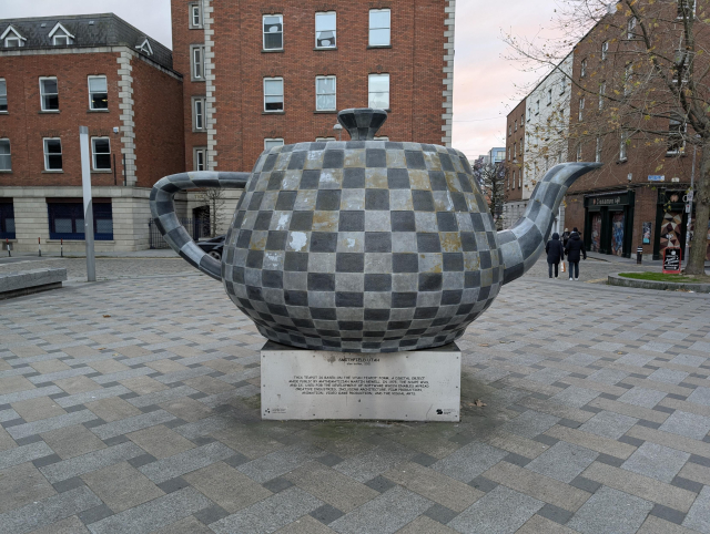 A large sculpture of the Utah teapot, with a checker board pattern. It stands on a pedestal in an open square with a similar checkered brick tile pattern, with orange brick buildings in the background.
