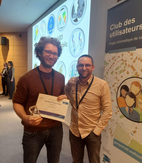 Joost Schouppe is standing indoors, holding a clear glass award in his right hand and a certificate labeled "GeoSpatial Award 2024 - Impact Award" in his left hand. He is wearing a dark sweater and a lanyard around his neck. Beside him is another person wearing a light-colored shirt, also with a lanyard. Behind them is a large screen displaying circular images related to geospatial themes and a banner with the text "Club des utilisateurs" and "Projet informatique de cartographie." 