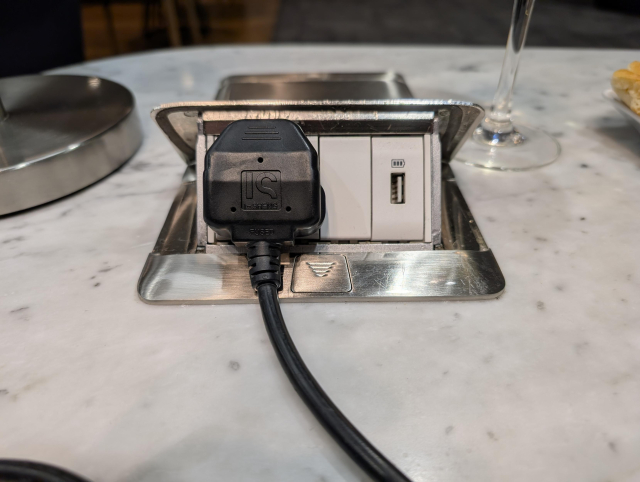 A black UK plug plugged into the socket. The captive cable leaving the bottom edge of the plug is fouling the flat surface of the table, making it impossible to plug in entirely and resulting in it not making electrical contact.