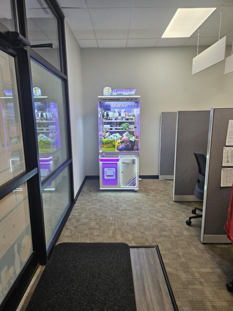 A big ole Claw Machine with purple and pink neon lights, very brightly lit on the otherwise beige and brown office space. It looks very out of place. 