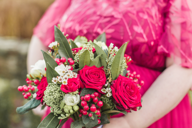 a close-up of a floral bouquet being held by a person wearing a vibrant pink dress. The bouquet consists of bright red roses, white flowers, and small red berries, accompanied by lush green leaves and some white buds. The vibrant colors of the flowers contrast beautifully with the pink fabric of the dress, which has a textured pattern that includes some floral or leaf-like designs. 