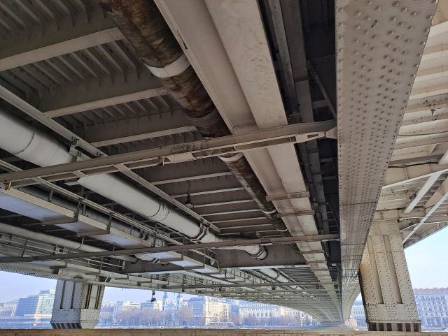 Photo of a large overarching metal structure, the underside of a bridge. There are pipes and bolts and beams. Between the pillars of rhe bridge, under the structure, the far shore of the Danube is visible with trees, tall buildings and a church.