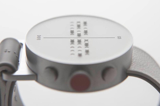 This is a close-up image of a Braille watch designed for visually impaired users. The circular watch face features raised Braille dots that allow the wearer to read the time by touch. The dots are arranged in a clear pattern, divided by a central line, likely indicating hours on one side and minutes on the other. The watch has a minimalist metal body, textured side buttons, and a leather strap visible in the background. Its design is sleek and functional, emphasizing accessibility.