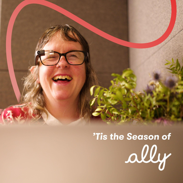 A woman with long, light brown hair is smiling brightly while wearing Envision Glasses. She is dressed in a festive red sweater with white holiday patterns, sitting in a cozy setting surrounded by green plants. The image has a warm holiday vibe with a soft pink decorative swirl in the background. The text reads, "'Tis the Season of Ally," highlighting the theme of accessibility and festive cheer.