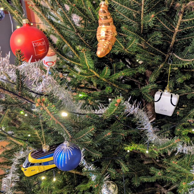 A detail of a decorated Christmas tree. On top left, a red ball with logo of Oskar is hanging. Behind it hangs another ball in the shape of a paper coffee cup filled with cappuccino having Kiosk logo on it.
There's also a croissant shaped decoration hanging in the middle and a ball shaped like a pasta box from Julia's bearing the logo on the right. 
A ball shaped like NS ICNG train hangs in the bottom left together with an ordinary blue ball.