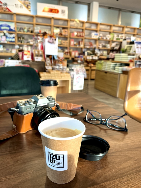 Coffe in a paper cup on a table with a camera and glasses. Blurred background of a book store inside a coffee shop