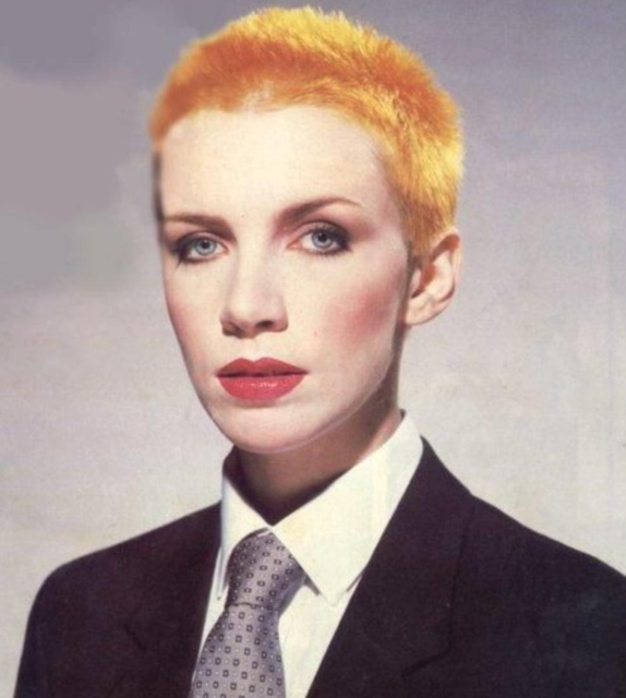 Photo of a young Annie Lennox wearing a business suit and tie with her short pixie hair dyed orange.