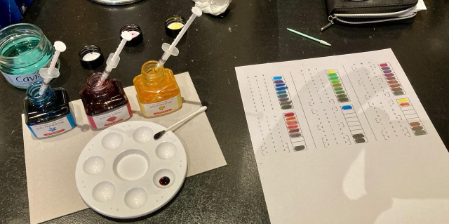 Photo of a desk with a three bottles on ink on the left, blue, red, and yellow, each with a small syringe in it. In the foreground is a small paint mixing palette. On the right is a sheet of paper with many colour samples, each corresponding to a different ratio of the three inks.