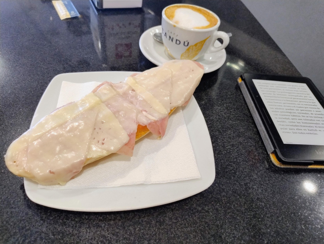 Foto de tostada de tomate, york y queso fundido, café con leche y Kindle , todo en una mesa de bar