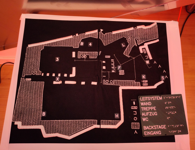 Blick auf einen quadratischen Tisch, von oben. Auf dem Tisch ist ein taktiler Plan des Erdgeschosses des CCH festgemacht.
Der Plan besteht aus vier Stück schwarzem Stoff, die zusammen genäht wurden. Der Grundriss des Stockwerks wurde mit weißem Garn eingestickt. Zusätzlich wurden 3D-gedruckte Plättchen angenäht, die einzelne Buchstaben oder Ziffern in Profilschrift enthalten. Rechts unten erklärt eine Legende die verschiedenen Stick-Stiche in Profilschrift und Braille.