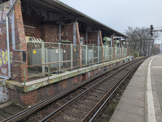 A view of a substation with 5 big high village transformers. In front of them there's a railway electrified by a third rail partially covered from top and bottom.