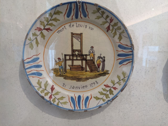 un plato de cerámica que data de la revolución francesa, con decoraciones a color y un motivo central de la decapitación de Luis XVI que lo muestra al pie de la guillotina con el cura, el verdugo y una persona con una espada y una antorcha.