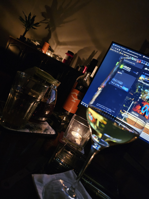 v popredí pohár vína za ním monitor kde je aktualizovaná stránka mastodonu, za monitorom fľaša vína, vedľa pred ňou čaj v sklenenej kanvici s pohárom vedľa, v pozadí pri stene polica kde sviečka vrhá tieň na bielu stenu, taká malá palma, na polici je random kade čo 