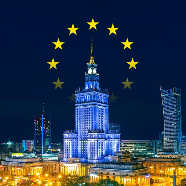 A photo of the Palace of Culture and Science in Warsaw, Poland, illuminated at night with the European Union flag superimposed.