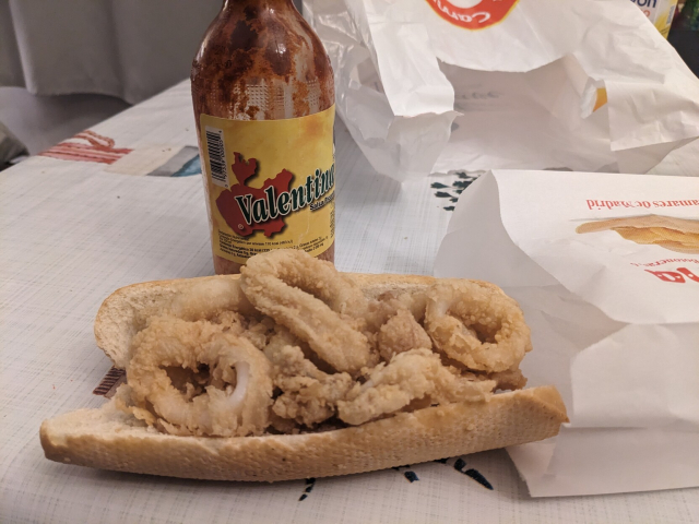 un bocadillo de calamar (un pan a la mitad de ser un bolillo o una baguette relleno de calamares en tempura) en primer plano y una salsa Valentina detrás