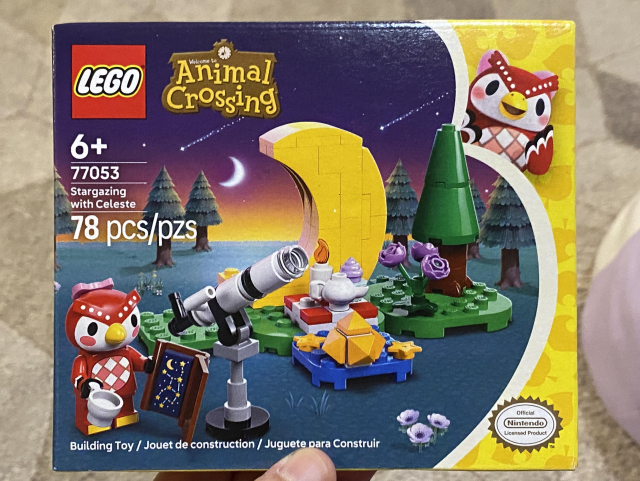 Box cover art for the Celeste LEGO Animal Crossing set showing Celeste, a red owl, holding a teacup and a star chart book, standing by a telescope. In the background is a grassy area with a large yellow crescent moon and a picnic table with a candle, a teapot, and a purple cupcake. I’m from of the table is a blue area with 2 stars and a yellow crystal on it. Further in the background is an evergreen tree and a trio of purple roses. 
