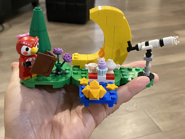 A hand is holding the whole, assembled LEGO set with all the items and background described for the box art picture, and Celeste holding the star chart book standing by the evergreen tree and roses. 